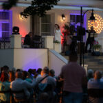 Na scenie przed białym budynkiem artystka śpiewa przy muzykach. Publiczność siedzi na krzesłach. Kolorowe światła oświetlają występ.