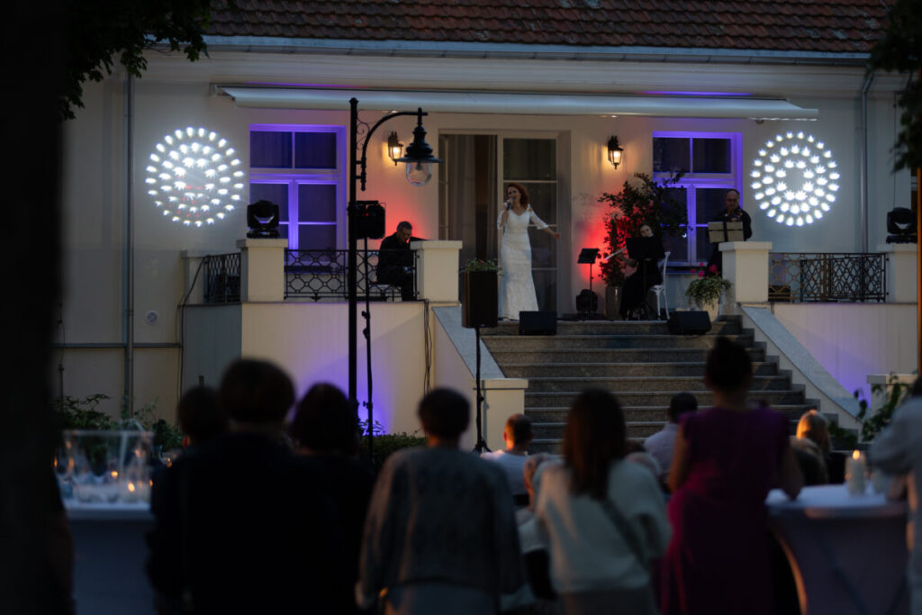 Na zdjęciu jest koncert na schodach przed jasnym budynkiem z czerwonymi dachówkami. Scena jest oświetlona białymi i fioletowymi światłami. Widownia stoi przed sceną w cienistym oświetleniu.