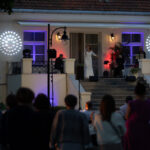 Na zdjęciu jest koncert na schodach przed jasnym budynkiem z czerwonymi dachówkami. Scena jest oświetlona białymi i fioletowymi światłami. Widownia stoi przed sceną w cienistym oświetleniu.