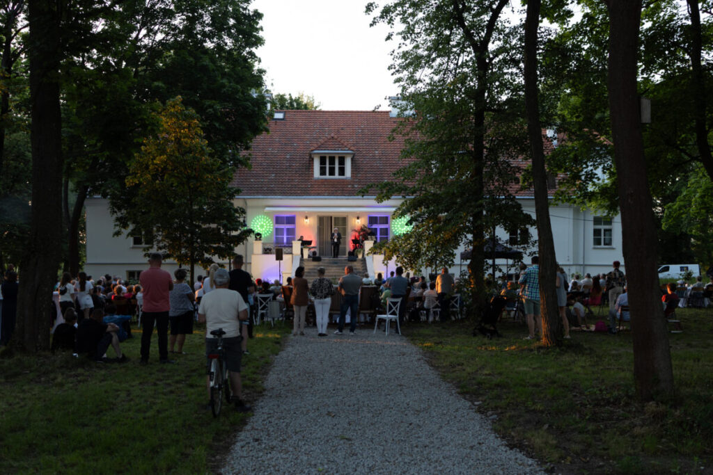 Publiczność słucha koncertu przed białym budynkiem w otoczeniu drzew. Scena oświetlona kolorowymi światłami. Wieczorny klimat.