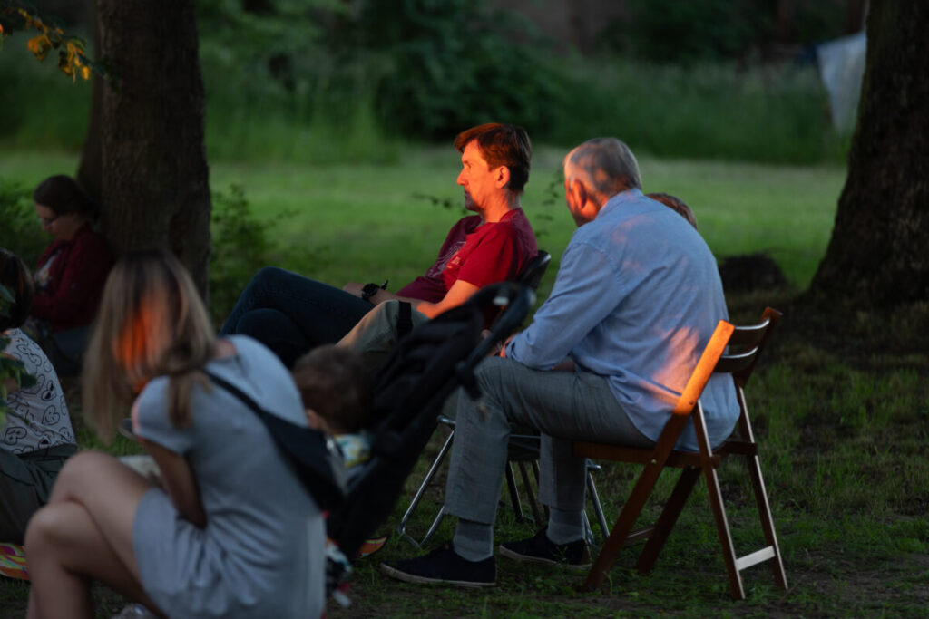 Grupa osób siedzi na krzesłach na trawie w parku. Tło jest zielone z drzewami, a światło zachodzącego słońca oświetla scenę. Dwie z osób intensywnie rozmawiają.