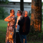 Na zdjęciu troje ludzi stoi obok drzewa podziwiając coś. Kobieta w sukience w kwiaty i dwaj mężczyźni, widoczne ciepłe światło zachodu. W tle zielona trawa i rozmyte drzewa.