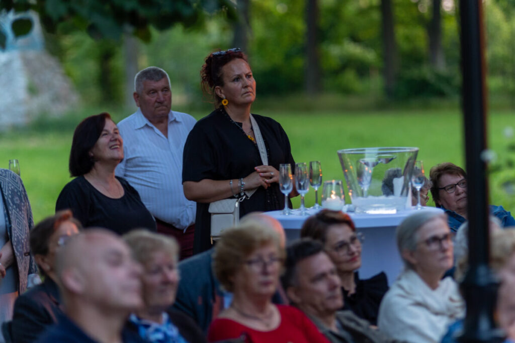Na zdjęciu grupa ludzi na otwartej przestrzeni. W tle zielone drzewa, na stole kieliszki i wiaderko z lodem. Jedna osoba stoi, reszta siedzi.