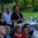 Na zdjęciu grupa ludzi na otwartej przestrzeni. W tle zielone drzewa, na stole kieliszki i wiaderko z lodem. Jedna osoba stoi, reszta siedzi.