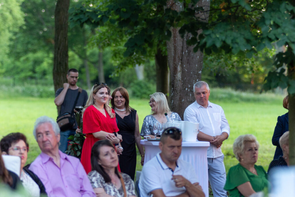 Na zdjęciu grupa ludzi na świeżym powietrzu. Osoby stoją przy stole wśród zieleni drzew. W tle widać zrelaksowaną atmosferę.