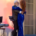 Mężczyzna w ciemnym garniturze i kobieta w błyszczącej niebieskiej sukni tańczą na tle jasnej ściany. Obok stoi keyboard. Romantyczne ujęcie z latarnią.