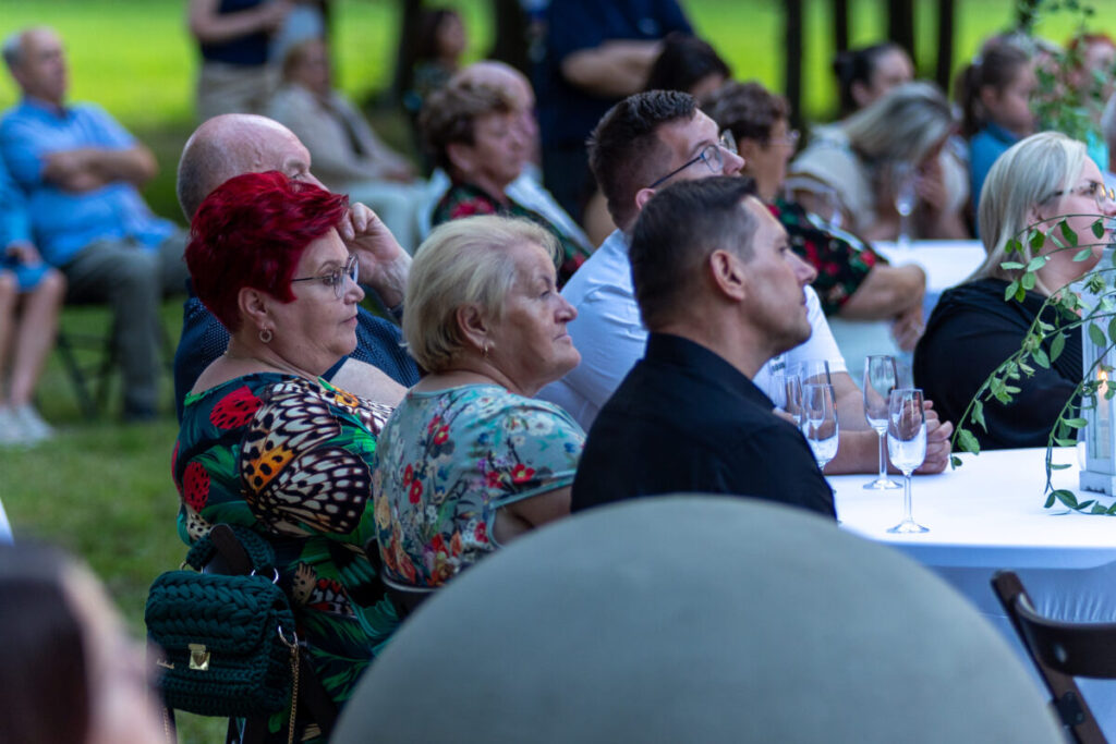 Na zdjęciu widać grupę ludzi przy stole, na zewnątrz na zielonej trawie. Osoby są odwrócone, zwracają uwagę na coś poza kadrem. Kolory są naturalne, dominuje zieleń i różnorodne kolory ubrań.