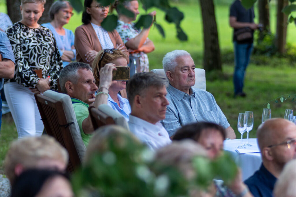 Grupa ludzi siedzi i stoi na świeżym powietrzu przy stole, w otoczeniu zielonego parku. Niektórzy trzymają telefony, a na stole są kieliszki. Atmosfera jest spokojna, a tło stanowią drzewa.