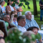 Grupa ludzi siedzi i stoi na świeżym powietrzu przy stole, w otoczeniu zielonego parku. Niektórzy trzymają telefony, a na stole są kieliszki. Atmosfera jest spokojna, a tło stanowią drzewa.