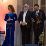 Troje ludzi stoi na scenie. Kobieta w niebieskiej sukni, mężczyźni w eleganckich garniturach, jeden trzyma róże. Tło ma ciepłe odcienie.