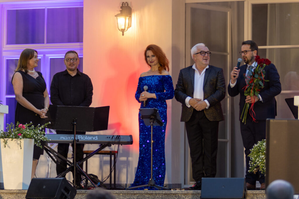 Na zdjęciu grupa elegancko ubranych osób na scenie. W tle fioletowe i czerwone oświetlenie. Obok nich stoi fortepian i mikrofon.