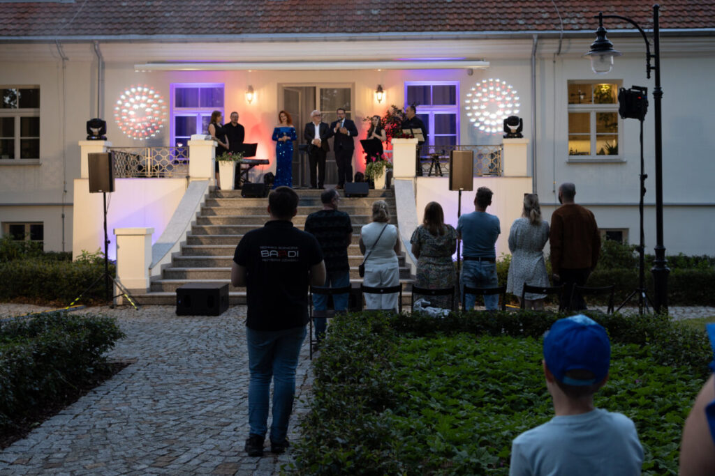 Na zdjęciu jest wieczorna uroczystość przed budynkiem z czerwonym dachem. Grupa ludzi stoi na schodach sceny, podświetlona niebieskim i fioletowym światłem. Widzowie oglądają wydarzenie na kamiennej ścieżce otoczonej zielenią