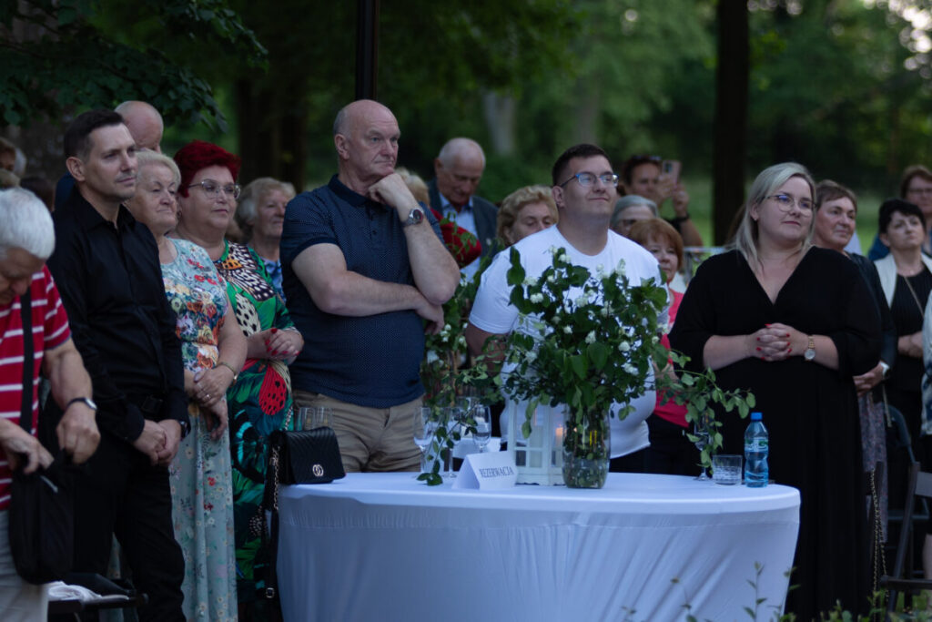 Grupa ludzi stoi na zewnątrz podczas wydarzenia. Na pierwszym planie stół z zielonymi roślinami i białym obrusem. W tle zielone drzewa.