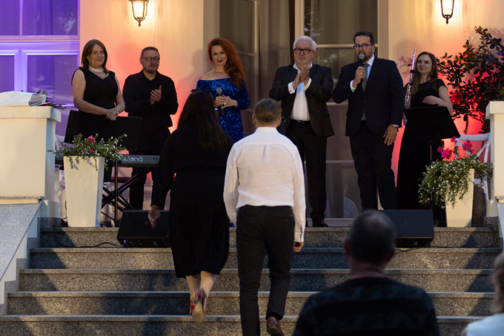 Grupa ludzi stoi na schodach przed budynkiem oświetlonym na fioletowo i pomarańczowo. Na przodzie, para idzie w ich stronę. W tle rośliny i instrumenty.