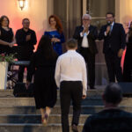 Grupa ludzi stoi na schodach przed budynkiem oświetlonym na fioletowo i pomarańczowo. Na przodzie, para idzie w ich stronę. W tle rośliny i instrumenty.