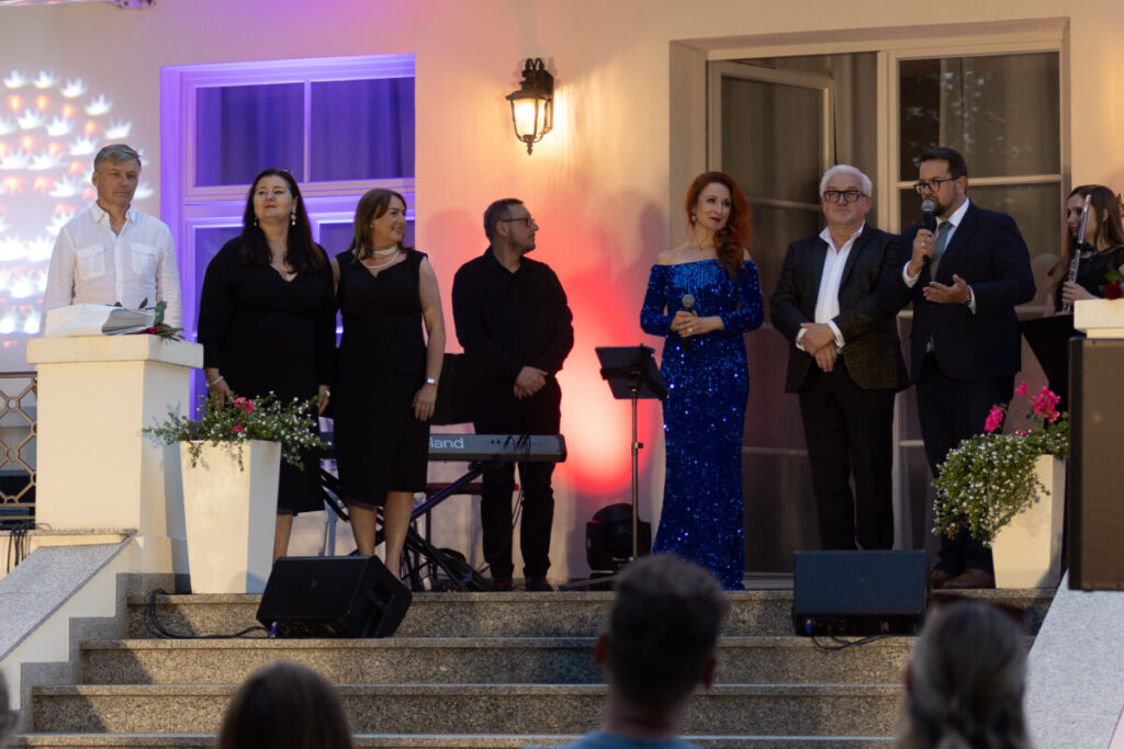 Grupa osób stoi na schodach w eleganckich strojach, w tle kolorowe światła i kwiaty. Atmosfera jest uroczysta.