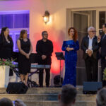 Na zdjęciu grupa ludzi na schodach, kobieta w niebieskiej sukni. Tło to budynek z lampką. Kwiaty w białych donicach po bokach.