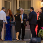 Grupa elegancko ubranych osób na scenie przed budynkiem. Kobieta w niebieskiej sukni trzyma różę. Tło oświetlone lampami, rośliny wokół.