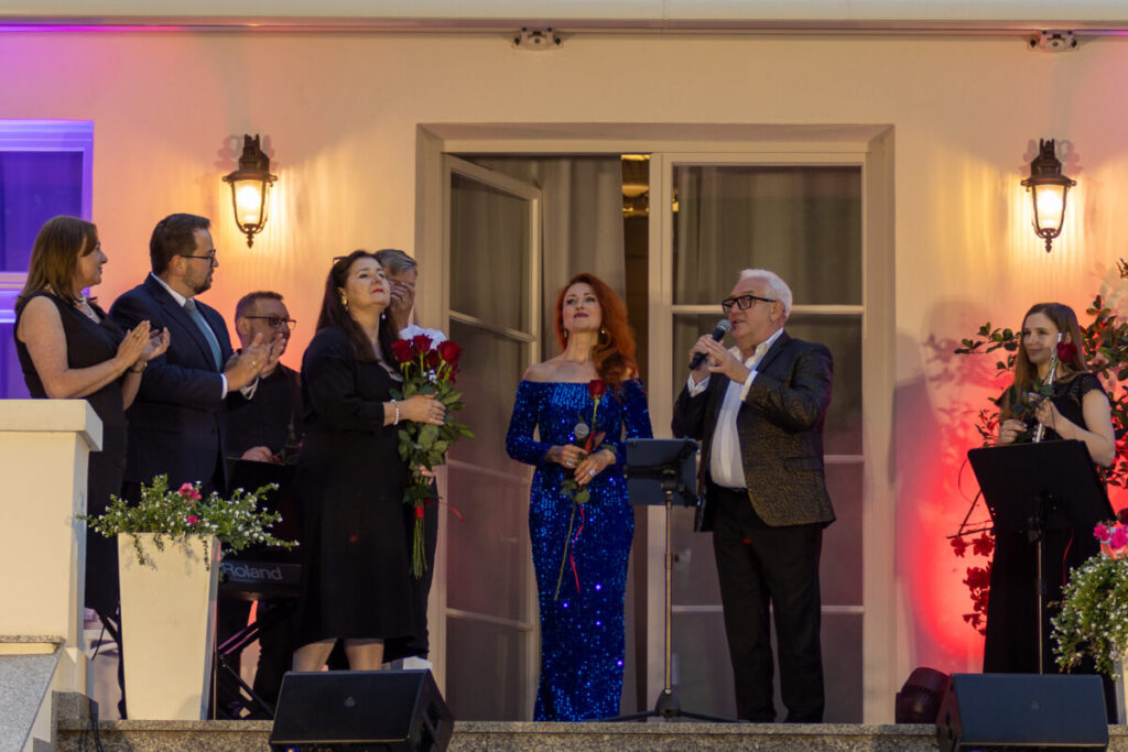 Grupa osób na scenie, elegancko ubrani, w centrum kobieta w niebieskiej sukni. Tło jasne z ciepłym oświetleniem, kwiaty w donicach.