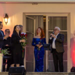 Grupa osób na scenie, elegancko ubrani, w centrum kobieta w niebieskiej sukni. Tło jasne z ciepłym oświetleniem, kwiaty w donicach.