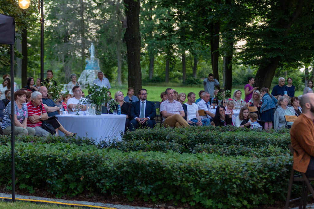 Ludzie siedzą w ogrodzie, oglądają wydarzenie. Naturalne oświetlenie, zielone tło drzew. Stół z białym obrusem i roślinami.