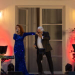 Na zdjęciu kobieta w niebieskiej sukience śpiewa z mężczyzną w garniturze. Tło jest kremowe z lampami i roślinami. Światła są ciepłe, w odcieniach czerwieni.