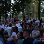 Na zdjęciu widoczna jest grupa ludzi siedzących przy okrągłych stolikach na zewnątrz, otoczonych drzewami. Jest wieczór, oświetlenie dają lampy i świeczki. Uczestnicy zwróceni są w jednym kierunku, jakby obser