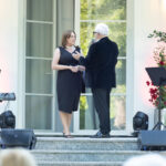 Dwoje ludzi stoi na schodach przed jasnymi drzwiami. Kobieta ma czarną sukienkę, a mężczyzna czarny garnitur. Obok nich kwiaty i sprzęt grający.