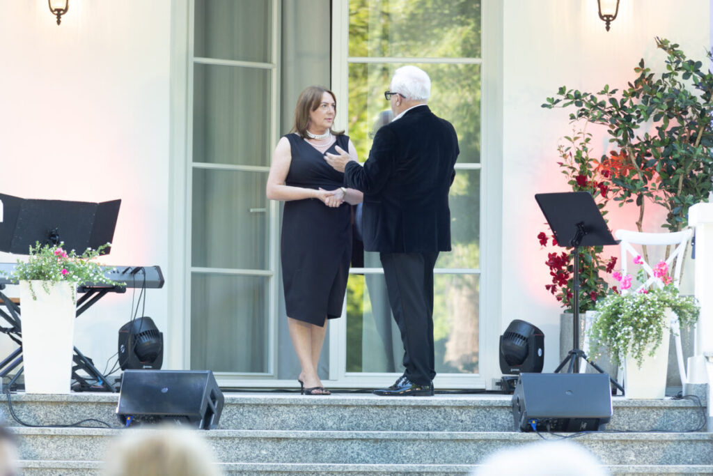 Para rozmawia na schodach w eleganckich strojach. Tło to jasna fasada z oknem i lampami. Obok stoją rośliny i instrumenty muzyczne.