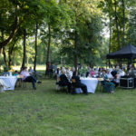 Grupa ludzi siedzi przy białych stołach na trawie w parku. Tło stanowią drzewa i zielona trawa. Obok widoczny jest czarny namiot.