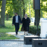 Para w eleganckich strojach spaceruje po parku. W tle zielone drzewa i trawnik. Dookoła widoczne elementy architektury.