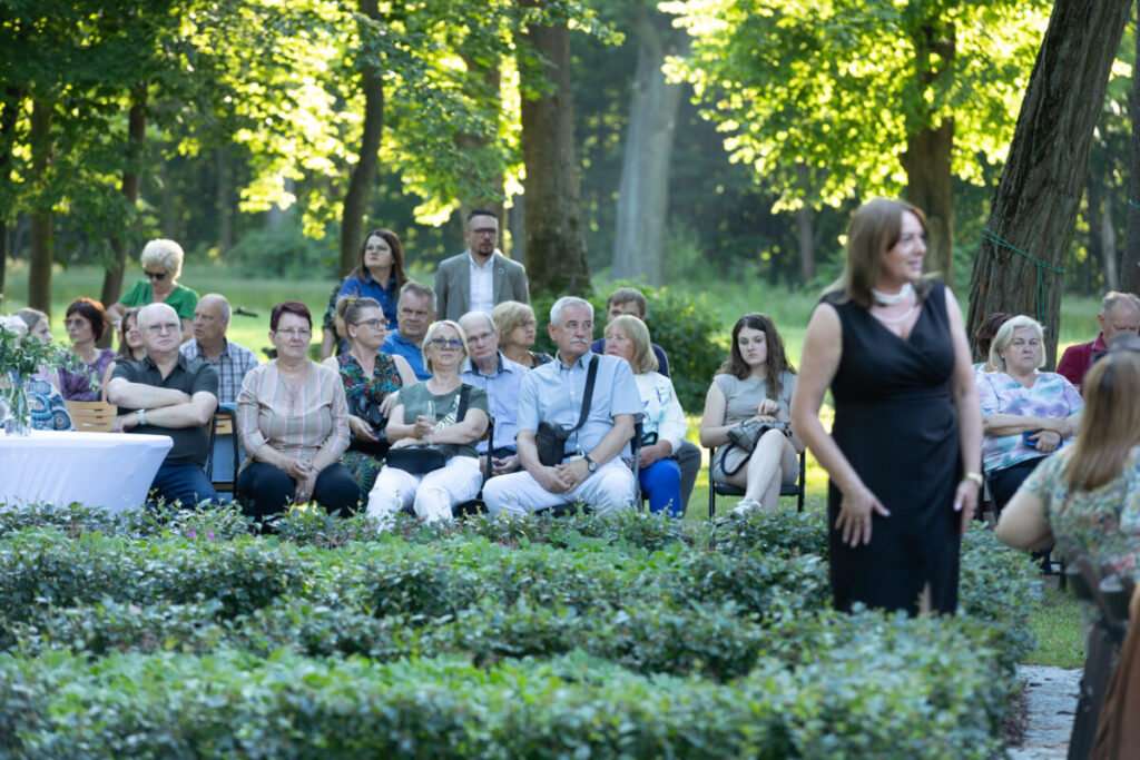 Grupa ludzi siedzi na ławkach w parku wśród zieleni drzew. Mężczyźni i kobiety różnego wieku obserwują coś przed sobą. Na pierwszym planie kobieta w czarnej sukni.