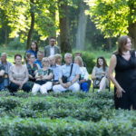 Grupa ludzi siedzi na ławkach w parku wśród zieleni drzew. Mężczyźni i kobiety różnego wieku obserwują coś przed sobą. Na pierwszym planie kobieta w czarnej sukni.