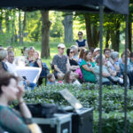 Grupa ludzi siedzi przy białych stolikach na zewnątrz w zielonym parku. Tło stanowią drzewa i krzewy. Sceneria jest jasna i słoneczna.