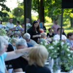 Zdjęcie przedstawia ludzi siedzących na zewnątrz latem. Tło to zielone drzewa i krzewy. Klimat spotkania jest spokojny.