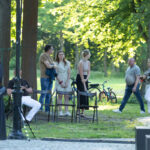 Grupa ludzi stoi na trawie w parku, otoczona drzewami. Widać rowery i krzesła. Tło jest zielone od liści drzew.