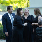 Grupa pięciu osób, ubranych elegancko, rozmawia ze sobą w ogrodzie. Tło jest zielone, słoneczne. Atmosfera wygląda na uroczystą.