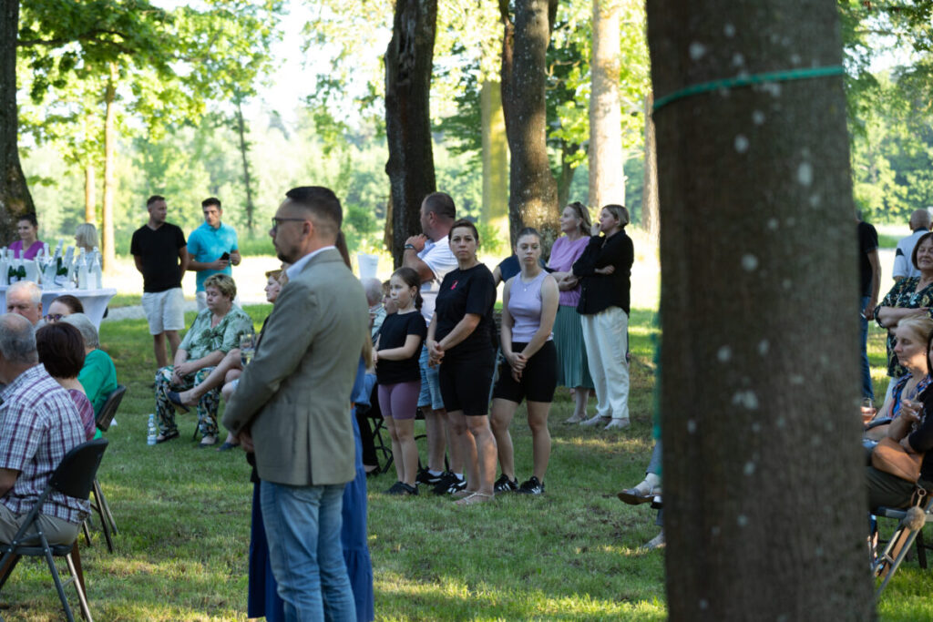 Grupa ludzi stoi i siedzi na zielonej trawie w lesie. W tle drzewa i stoliki z napojami. Kolory są naturalne i jasne.