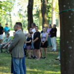 Grupa ludzi stoi i siedzi na zielonej trawie w lesie. W tle drzewa i stoliki z napojami. Kolory są naturalne i jasne.