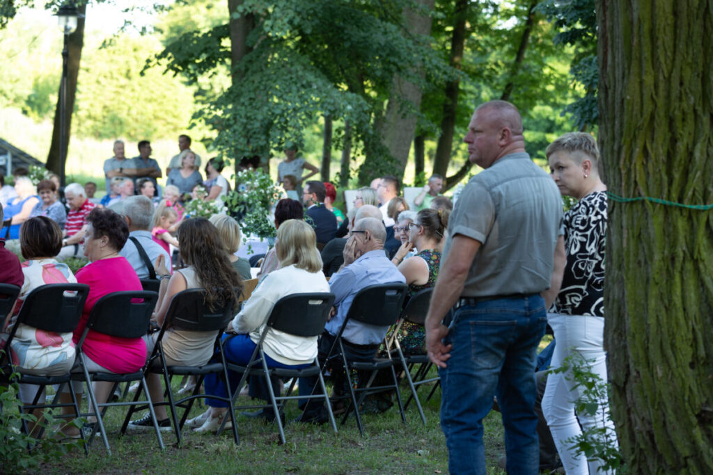 Zdjęcie przedstawia dużą grupę osób siedzących na krzesłach na zewnątrz. Tło stanowią zielone drzewa w parku. Dominują kolory zielony, czarny i szary.