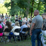 Zdjęcie przedstawia dużą grupę osób siedzących na krzesłach na zewnątrz. Tło stanowią zielone drzewa w parku. Dominują kolory zielony, czarny i szary.