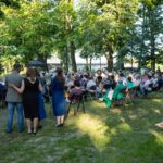 Grupa osób na pikniku w zielonym parku, siedzą na krzesłach. Słońce oświetla trawniki i drzewa. W tle biały budynek.