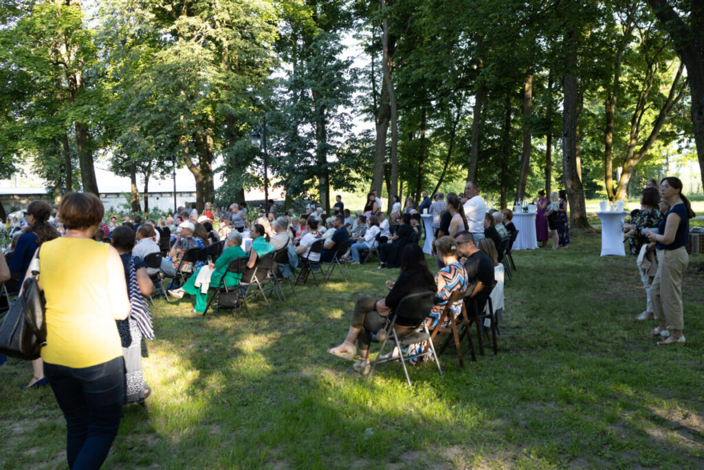 Na zdjęciu widać ludzi siedzących na krzesłach w parku. W tle widać drzewa i stoliki koktajlowe z białymi obrusami. Słońce rzuca cienie, a ludzie są w jasnych ubraniach.