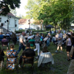 Na zdjęciu widoczna jest grupa ludzi siedzących na krzesłach w parku przed białym budynkiem. Tło stanowią zielone drzewa i trawnik. Po lewej stronie sceny widać oświetlenie i sprzęt do nagłośnienia.
