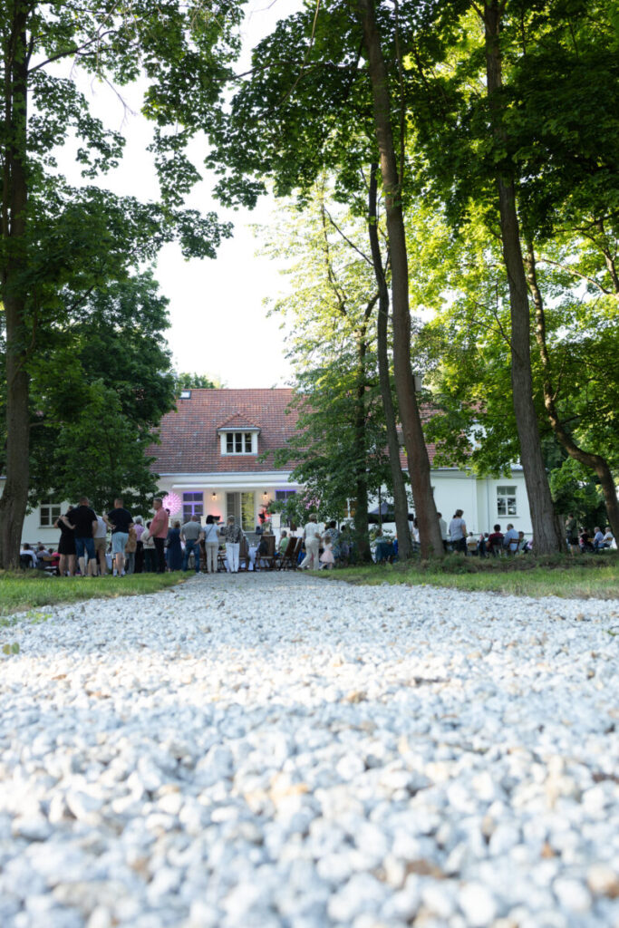 Ścieżka z białego żwiru prowadzi przez park do dużego, białego domu z czerwonym dachem. W tle zgromadzeni ludzie. Dookoła zielone drzewa.