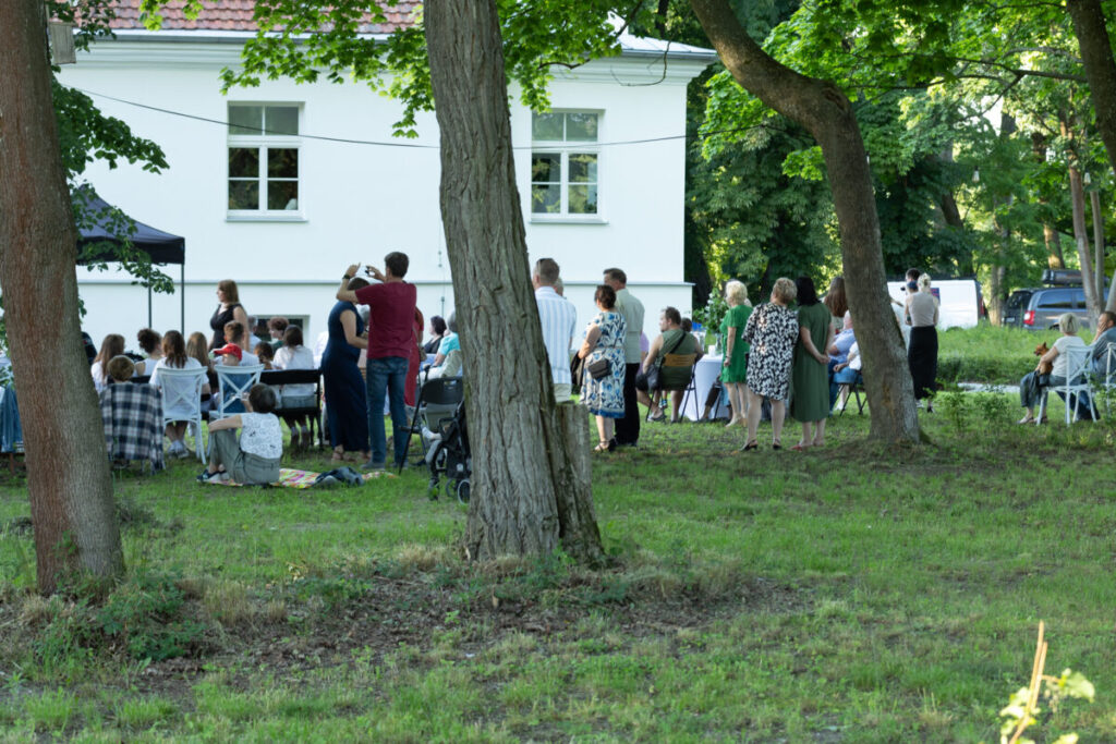 Plenerowy piknik przy białym budynku, zielony trawnik, drzewa. Ludzie na krzesłach i stojący, niektórzy robią zdjęcia.