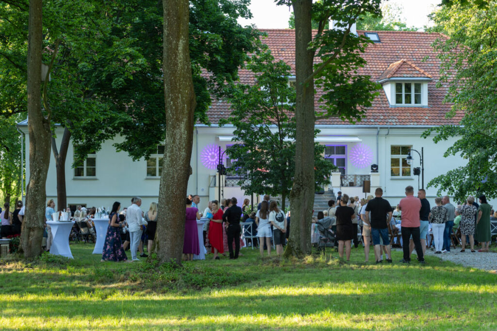 Spotkanie na zewnątrz przed białym budynkiem z czerwonym dachem. Goście stoją przy wysokich stolikach na trawie. Zielone drzewa otaczają przestrzeń.