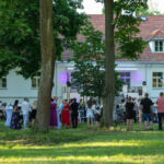 Spotkanie na zewnątrz przed białym budynkiem z czerwonym dachem. Goście stoją przy wysokich stolikach na trawie. Zielone drzewa otaczają przestrzeń.