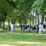 Grupa ludzi stoi na trawie w parku pełnym drzew. W tle widać biały budynek. Rowerki dziecięce są oparte o drzewa.