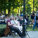 Grupa ludzi siedzących na krzesłach na tle zielonego parku. Przed nimi lampa, a w tle widać innych uczestników. Dominują zielenie i ciepłe kolory.
